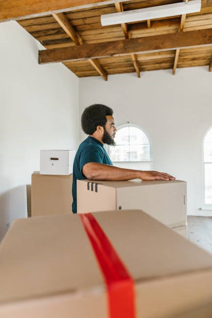 A man with a beard and curly hair, dressed in a blue shirt, is standing inside a spacious room with a high wooden ceiling and large arched windows, engaged in a home relocation process organized by St